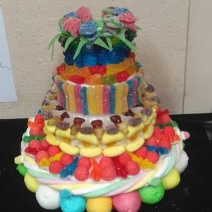 Peut inclure: Un gâteau de bonbons à plusieurs niveaux, composé d'une variété de bonbons colorés. Il présente des couches de bonbons gélifiés, de guimauves et d'autres sucreries, surmontées de fleurs en bonbons. La base est ornée de grosses boules de guimauve.