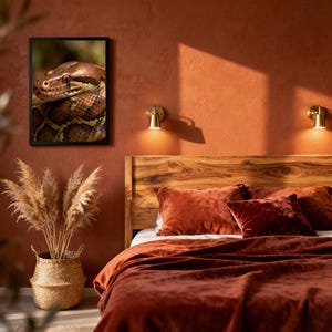 以下が含まれることがあります： 茶色とベージュの鱗を持つヘビの写真が額縁に入っており、錆色の寝具と枕のあるベッドの上に飾られています。乾燥したパンパスグラスが入った織りのバスケットがベッドの近くにあります。部屋は暖かく居心地の良い雰囲気です。