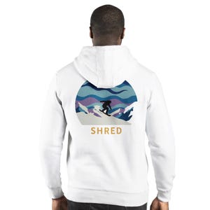 May include: White hoodie with a graphic of a snowboarder on a snowy mountain scene. The design features shades of blue, purple, and white, with the word "SHRED" in gold lettering below the image. A casual, sporty apparel item.