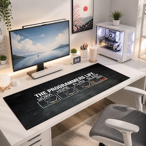 Può includere: Una moderna postazione di lavoro per computer con un ampio monitor che mostra una vista panoramica. Un tappetino da scrivania nero con la scritta "THE PROGRAMMERS LIFE" e un design a lista di controllo è di fronte al monitor. Un case per computer bianco e altri accessori completano lo spazio di lavoro.