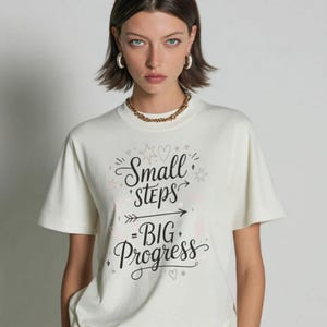 May include: Cream-colored t-shirt with the text "Small Steps - Big Progress" in a handwritten style. The shirt has short sleeves and a crew neck. The overall style is casual and comfortable.