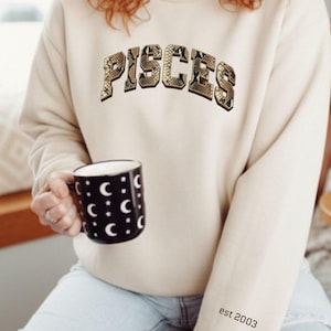 Könnte beinhalten: Beiges Sweatshirt mit dem Wort "PISCES" in einem Schlangenmuster auf der Brust. Der Ärmel hat den Text "est 2003". Eine schwarze Tasse mit weißen Monden wird in der Hand gehalten.