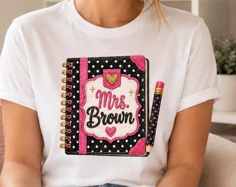 Könnte beinhalten: Ein weißes Kurzarm-T-Shirt mit einer Grafik eines schwarzen spiralgebundenen Notizbuchs mit weißen Punkten, rosa Akzenten und dem Text Mrs. Brown in Schreibschrift. Ein Bleistift mit rosa Radiergummi steckt an der Seite der Notizbuchgrafik.