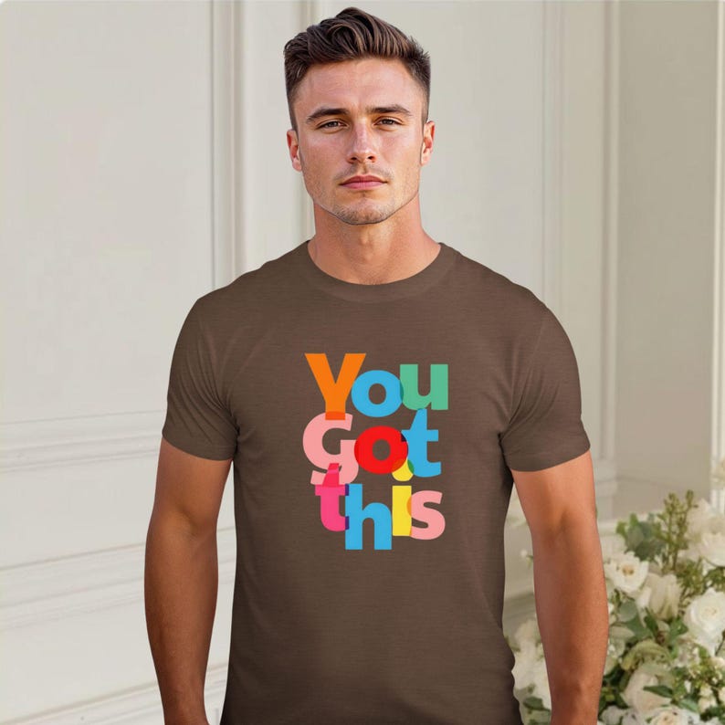 May include: A brown t-shirt featuring the phrase "You Got This" in vibrant, multi-coloured block letters. The text is stacked vertically, with each word in a different hue. The t-shirt has a classic crew neck design.