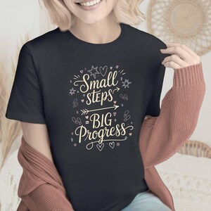 May include: A black t-shirt with the text "Small Steps Big Progress" in a decorative script. The design includes stars, hearts, and an arrow. The shirt is a dark color.