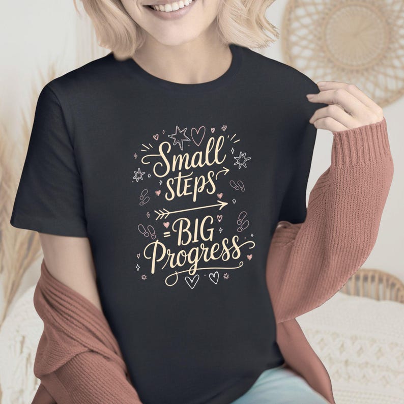May include: A black t-shirt with the text "Small Steps Big Progress" in a decorative script. The design includes stars, hearts, and an arrow. The shirt is a dark color.
