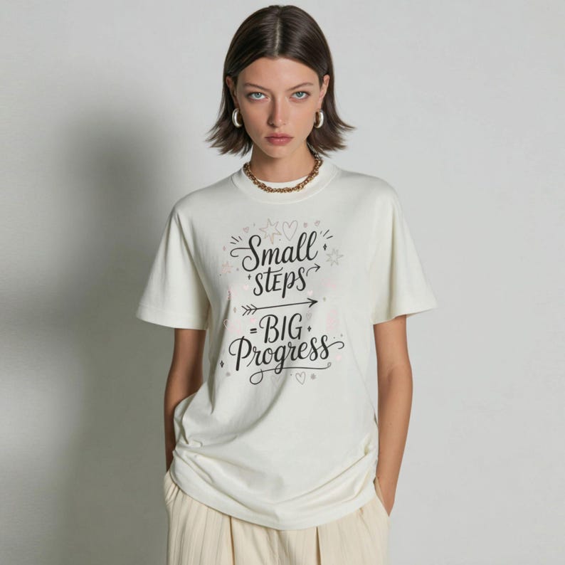 May include: Cream-colored t-shirt with the text "Small Steps - Big Progress" in a handwritten style. The shirt has short sleeves and a crew neck. The overall style is casual and comfortable.