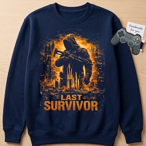 May include: Navy blue sweatshirt featuring a hooded figure holding a rifle against a fiery cityscape backdrop. The words "LAST SURVIVOR" are printed in orange below the graphic. A small, gray game controller is in the upper right corner.
