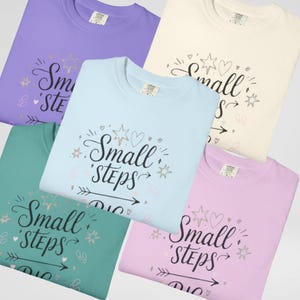 May include: A collection of five crew neck t-shirts in various pastel colors: lavender, cream, light blue, teal, and pink. Each shirt features the phrase "Small Steps" in a decorative script with star and heart accents.