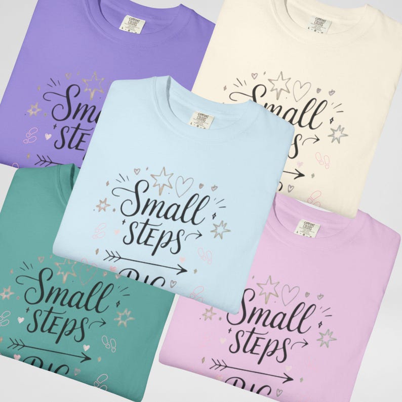 May include: A collection of five crew neck t-shirts in various pastel colors: lavender, cream, light blue, teal, and pink. Each shirt features the phrase "Small Steps" in a decorative script with star and heart accents.