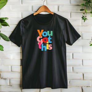May include: Black t-shirt with the phrase "You Got This" in colourful, stacked letters. The text features orange, green, pink, red, blue, and yellow hues. The shirt is hanging on a wooden hanger against a white brick wall.
