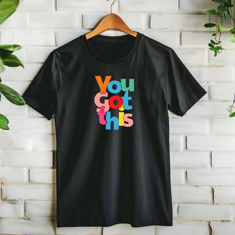 May include: Black t-shirt with the phrase "You Got This" in colourful, stacked letters. The text features orange, green, pink, red, blue, and yellow hues. The shirt is hanging on a wooden hanger against a white brick wall.