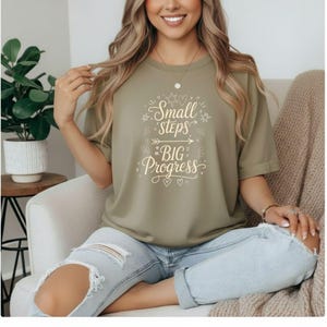 May include: Olive green t-shirt with the text "Small Steps Big Progress" in a white, whimsical font. The shirt features a crew neck and short sleeves. The design includes stars, arrows, and hearts.