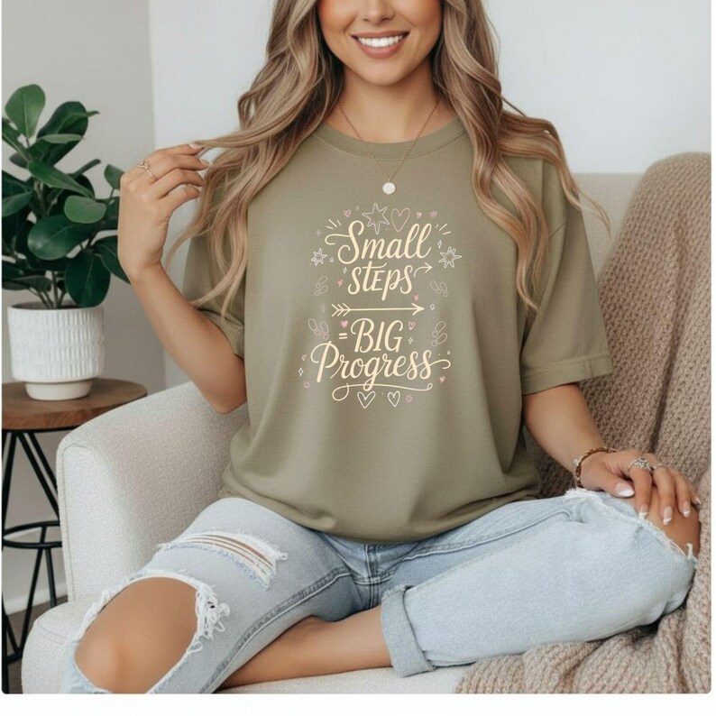 May include: Olive green t-shirt with the text "Small Steps Big Progress" in a white, whimsical font. The shirt features a crew neck and short sleeves. The design includes stars, arrows, and hearts.