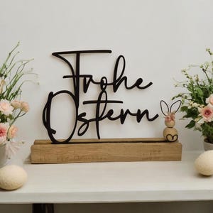 Puede incluir: Decoración de Pascua de madera con la frase alemana "Frohe Ostern" en escritura negra. El letrero se asienta sobre una base de madera, con una pequeña figura de conejito. Flores y huevos decorativos flanquean el letrero.