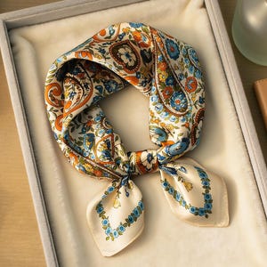 May include: A cream-colored silk scarf with a paisley and floral pattern in shades of orange, blue, and green. The scarf is folded and tied, displayed in a gray box with a soft, white interior. The scarf is a square shape.