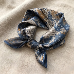 Puede incluir: Un pañuelo cuadrado de seda con un estampado repetitivo en tonos azules, marrones y beige. El pañuelo está anudado en el centro, con los extremos extendidos. La tela tiene un brillo sutil.
