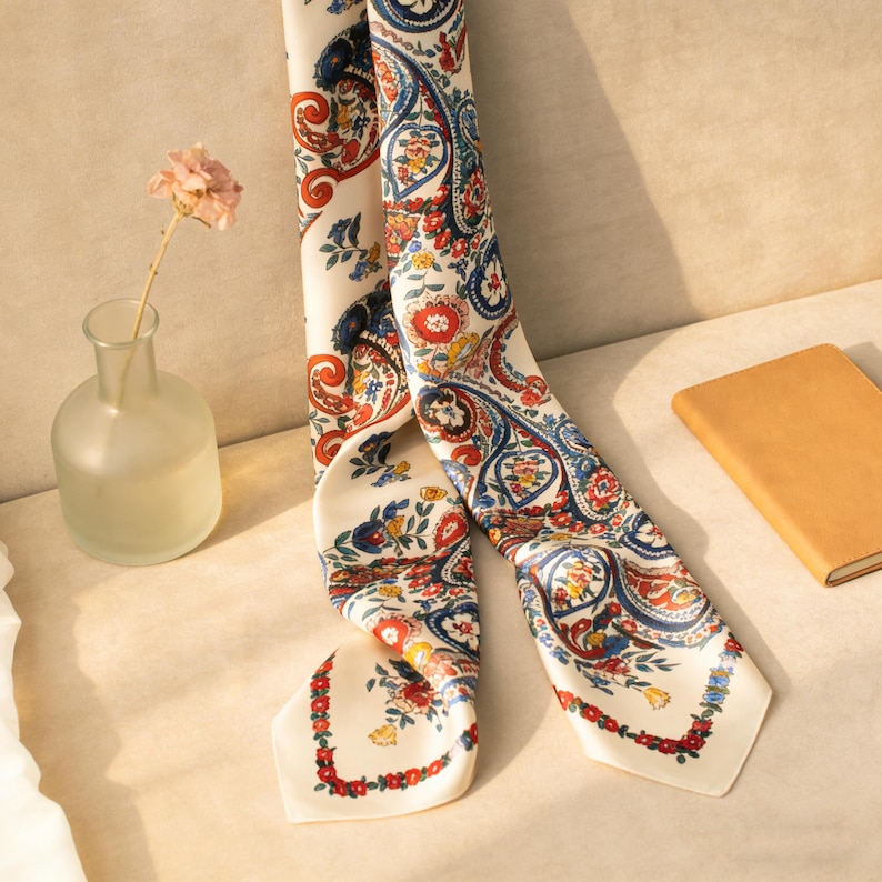 May include: A cream-colored silk scarf with a paisley pattern in red, blue, and yellow. The scarf is draped over a surface, with a small vase holding a dried flower and a tan notebook in the background.
