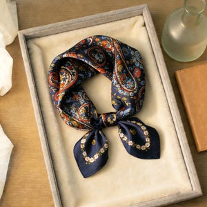 May include: A navy blue silk scarf with a paisley pattern in shades of orange, blue, and white. The scarf is square and folded, with a floral border at the ends. It is displayed in a gray velvet-lined tray.