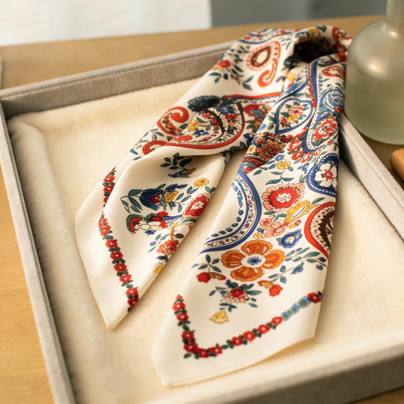 May include: Ivory-colored silk scarf with a vibrant floral and paisley pattern in red, blue, and orange. The scarf is folded and displayed in a gray tray, showcasing its intricate design and color palette. A decorative accessory.
