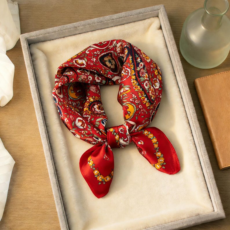 May include: A red silk scarf with a paisley pattern and floral accents. The scarf is knotted and displayed in a gray velvet-lined box. The scarf's design includes yellow, white, and navy blue details.