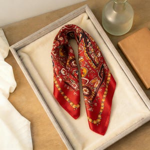 May include: A red silk scarf with a paisley pattern in yellow, black, and white. The scarf is folded and displayed in a gray tray with a cream-colored lining. A small glass vase and a tan leather notebook are in the background.
