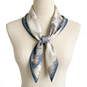 May include: A square silk scarf with a floral pattern in cream, blue, and brown tones. The scarf is tied around a mannequin's neck, showcasing its design and the navy blue border. The scarf's design features detailed floral illustrations.