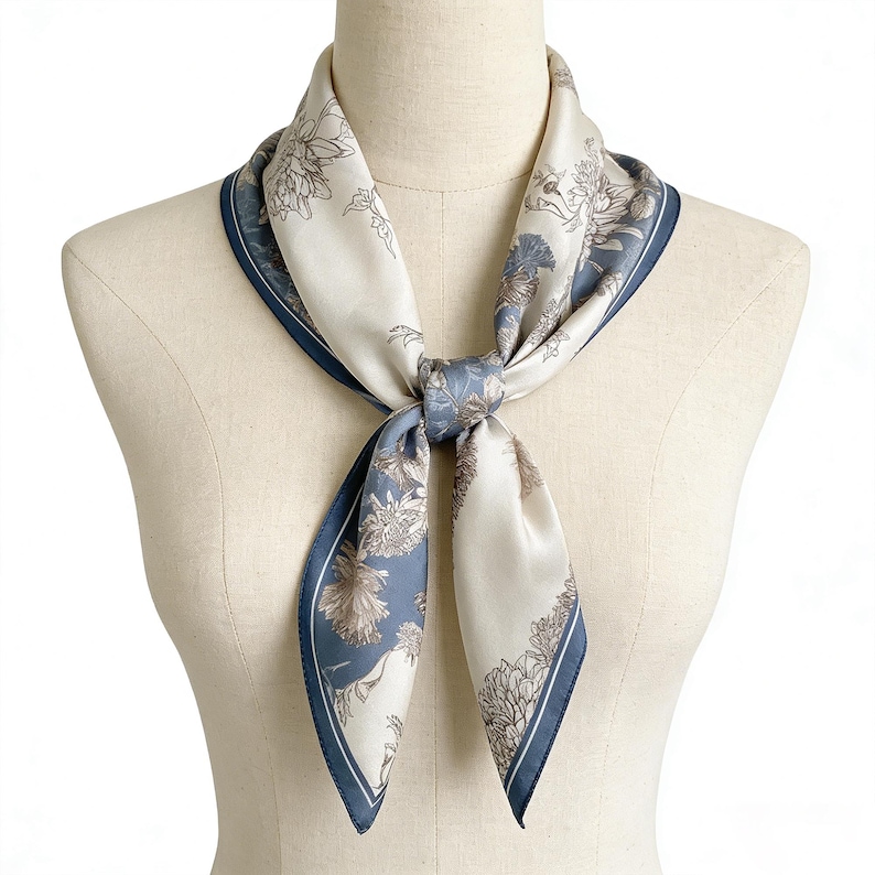 May include: A square silk scarf with a floral pattern in cream, blue, and brown tones. The scarf is tied around a mannequin's neck, showcasing its design and the navy blue border. The scarf's design features detailed floral illustrations.