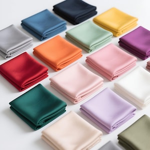 Może przedstawiać: Kolekcja złożonych kwadratowych poszetek jedwabnych w różnych kolorach, w tym granatowym, niebieskim, pomarańczowym, zielonym, różowym i fioletowym. Poszetki są starannie ułożone, prezentując gładką fakturę i żywe odcienie. Idealne do formalnych strojów.