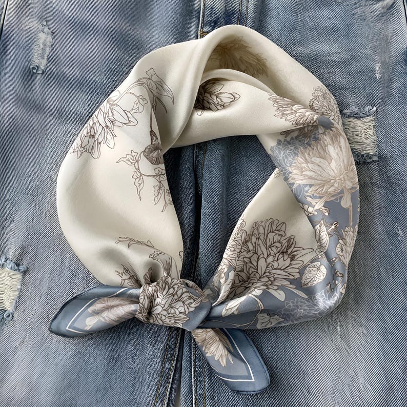 May include: A square silk scarf featuring a floral design in white, grey, and blue hues. The scarf is knotted, highlighting the intricate botanical pattern. The backdrop is a light blue denim fabric.