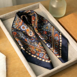 May include: A navy blue silk scarf with a paisley pattern in orange, blue, and white. The scarf is folded and displayed in a gray tray, with a leather wallet and a glass bottle in the background.