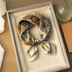 May include: A cream-colored silk scarf with a paisley and floral pattern in shades of orange, blue, and green. The scarf is folded and tied, displayed in a gray box with a soft, white interior. A decorative accessory.