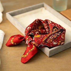 May include: A red silk scarf with a paisley pattern and floral accents. The scarf is knotted and lies in a gray tray on a wooden surface. The scarf's design includes yellow, blue, and white details.