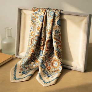 May include: A cream-colored silk scarf with a paisley pattern in shades of blue, orange, and brown. The scarf is draped over a gray display box. The edges of the scarf are trimmed with a blue floral border.