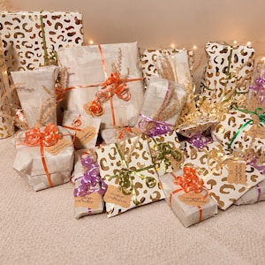 Peut inclure: Une collection de divers paquets cadeaux emballés dans du papier blanc à motif léopard doré ou du papier blanc uni. Les cadeaux sont noués avec des rubans colorés orange, verts et violets, et décorés de petites tiges florales séchées.