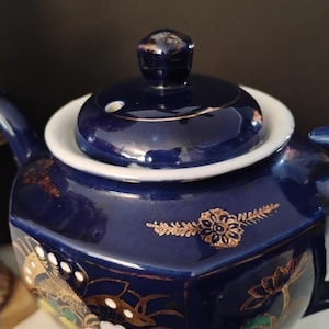 May include: A navy blue ceramic teapot with a lid and a handle. The teapot is decorated with gold floral designs and white, green, and yellow accents. The lid has a small knob and a white rim.
