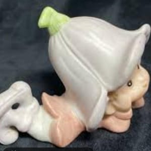 May include: A ceramic figurine of a small creature with a large white flower-shaped head. The flower has a green top and the creature has a light tan face and limbs. The figurine is in a reclining pose with pink accents.