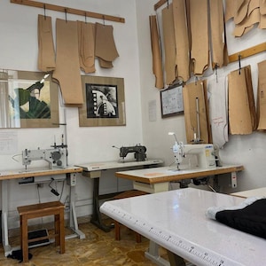 May include: A tailor's workshop with sewing machines, cutting tables, and patterns hanging on the wall. The room has a bright, well-lit atmosphere, with a wooden stool and framed artwork adding detail to the space.