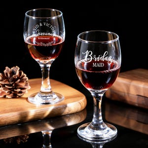May include: Two clear wine glasses filled with red liquid. One glass has the text "Vid & Victor Jessica Bridesmaid" etched on it, and the other says "Bridesmaid". The glasses are on a wooden surface with a pinecone.