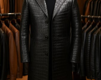 May include: A black, long leather coat with a crocodile skin texture is displayed on a mannequin. The coat has a classic design with a notched lapel, three buttons, and a long length. The mannequin is in a clothing store.