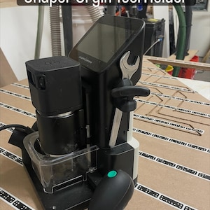 Puede incluir: Un soporte para herramientas Shaper Origin negro y plateado con una llave inglesa adjunta. La herramienta tiene una pantalla y un mando a distancia negro con un botón verde. El texto "Shaper Origin Tool Holder" está en la parte superior de la imagen.