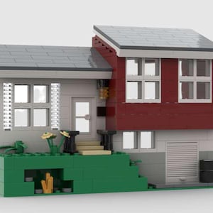 May include: A two-story house model constructed from colorful building blocks. The house features a gray roof, white walls, and a red upper level. Green blocks form a small yard with yellow flowers. A garage door and black barrels are visible.
