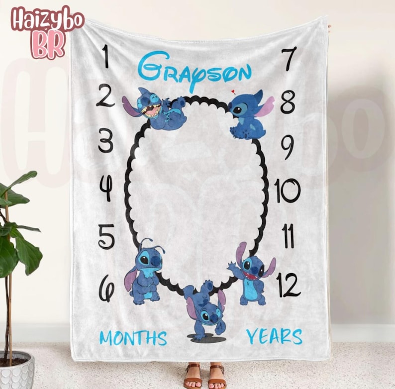 May include: A white milestone blanket featuring the name Grayson in blue text, surrounded by numbers 1 through 12, the words MONTHS and YEARS, and four blue Stitch characters arranged around a central black scalloped oval frame.