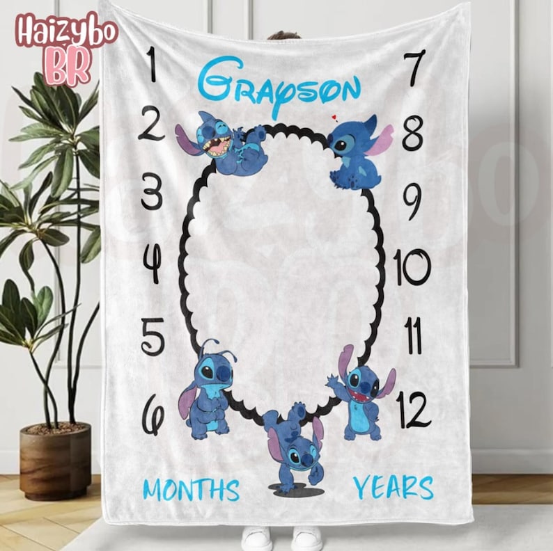 May include: A white milestone blanket featuring the name Grayson in blue text, numbers 1 through 12 in black, and illustrations of the blue character Stitch surrounding a central black oval frame, with the words MONTHS and YEARS printed in blue.