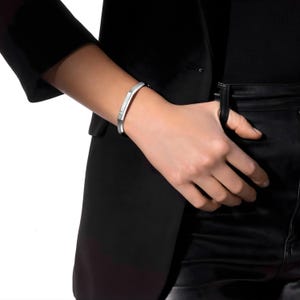 May include: A silver-colored bracelet with a geometric design. The bracelet is worn on a wrist, and the image shows a close-up view of the jewelry. The bracelet has a rectangular shape with a small, square accent.