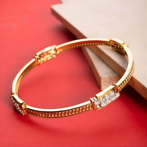 May include: A gold bangle bracelet with a hinged clasp and a series of small, sparkling stones. The bracelet has a textured design along the edges and is set against a red background.