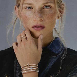 May include: Rose gold jewelry set featuring a ring and multiple bracelets. The ring has a floral design with small gemstones. The bracelets are adorned with gemstones and a circular emblem. The model is wearing a black leather jacket.