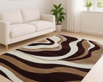 May include: A rectangular area rug with a wavy pattern in shades of brown, beige, and white. The rug is placed on a light-colored wooden floor in a living room setting. A white sofa and a framed picture are visible.