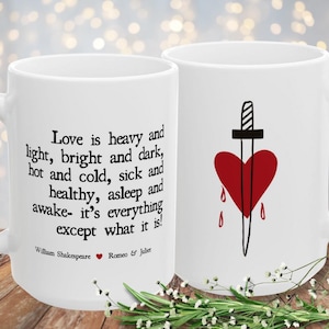 May include: Two white ceramic mugs. One mug features a quote from William Shakespeare's Romeo & Juliet: "Love is heavy and light..." The other mug has a red heart pierced by a dagger. Decorative floral accents are at the base.