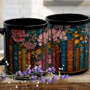 May include: Two black ceramic mugs with a floral and bookshelf design. The mugs feature pink flowers, colorful books, and a teal background. A sprig of lavender rests in front of the mugs on a wooden surface.